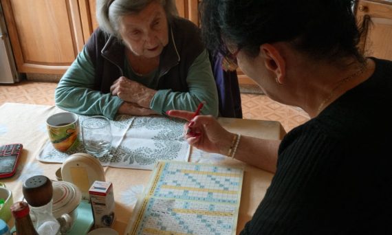 A senior woman and her caregiver work together on a crossword puzzle in a cozy kitchen.